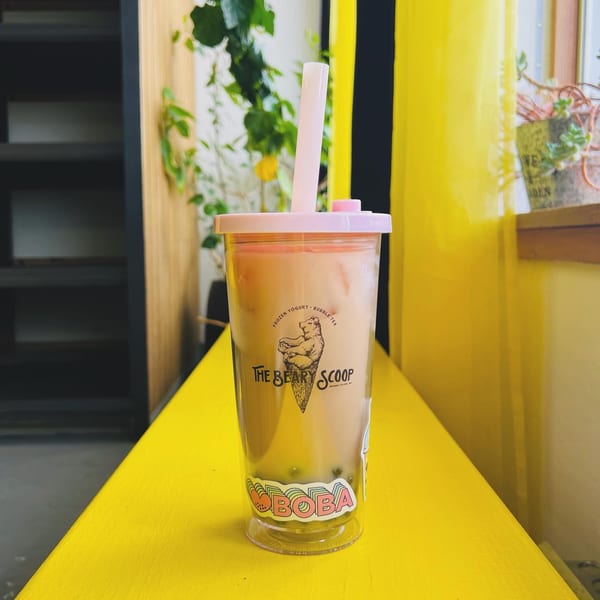 A Beary Scoop bubble tea with a pink lid and straw sits on a bright yellow surface.
