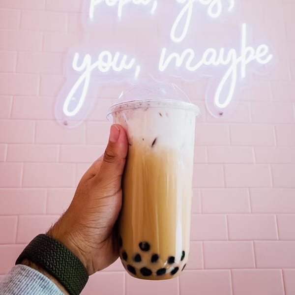 Hand holding a bubble tea with a pink tiled wall and neon sign in the background.