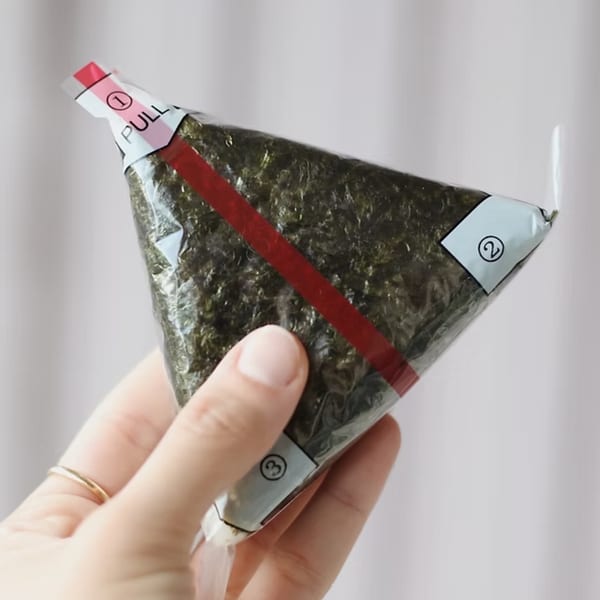 A hand holds a triangular onigiri wrapped in plastic with a red pull strip.
