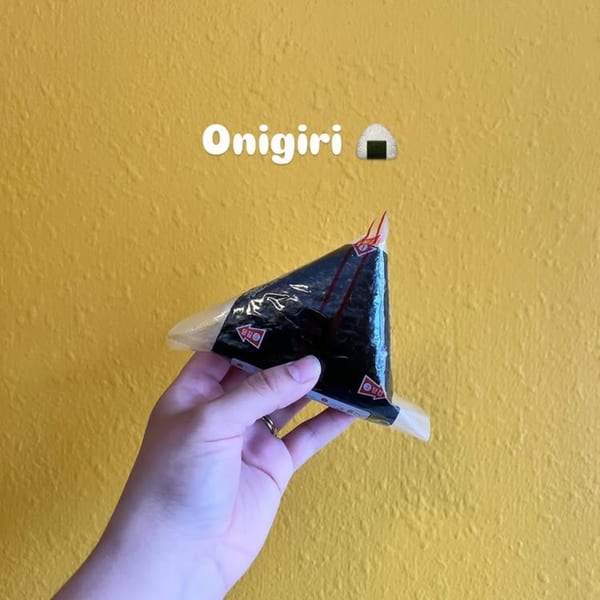 Hand holding a triangular, plastic-wrapped onigiri against a textured yellow wall with "Onigiri" text.